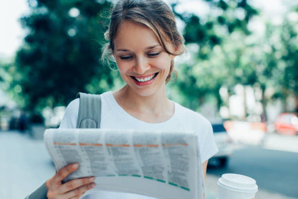Closeup portrait of smiling young beautiful woman holding drink and reading newspaper on street. Front view.