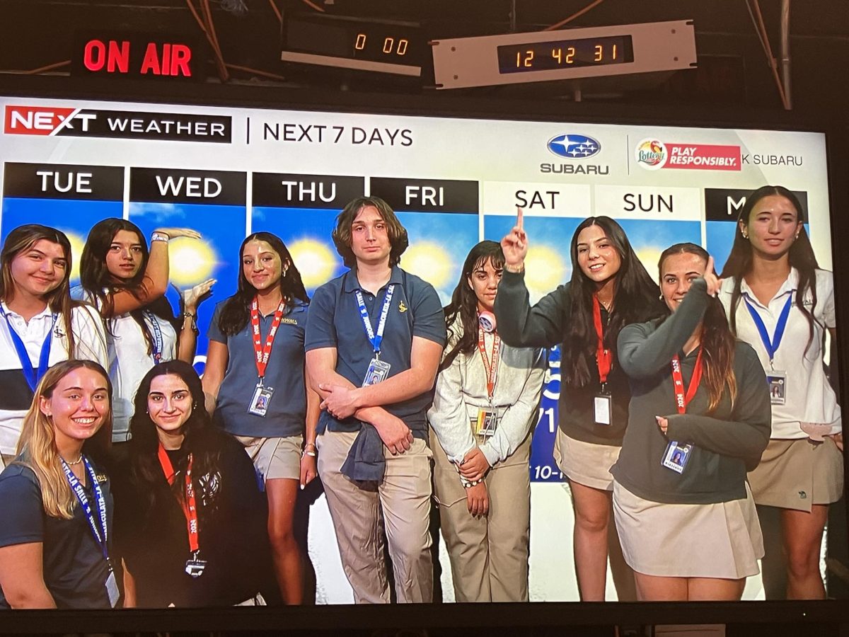 Brecelis Valdes, Daniella Coro, Ella Baker, Miguel Suarez Cabal, Veronica Mederos, Renata Alvarez Gil, Briana Del Oro, Valentina Lanza, and kneeling Gianna Ferrara and Eva de la Torre, pose in front of the green screen which has had the day's weather transposed for them in the background.