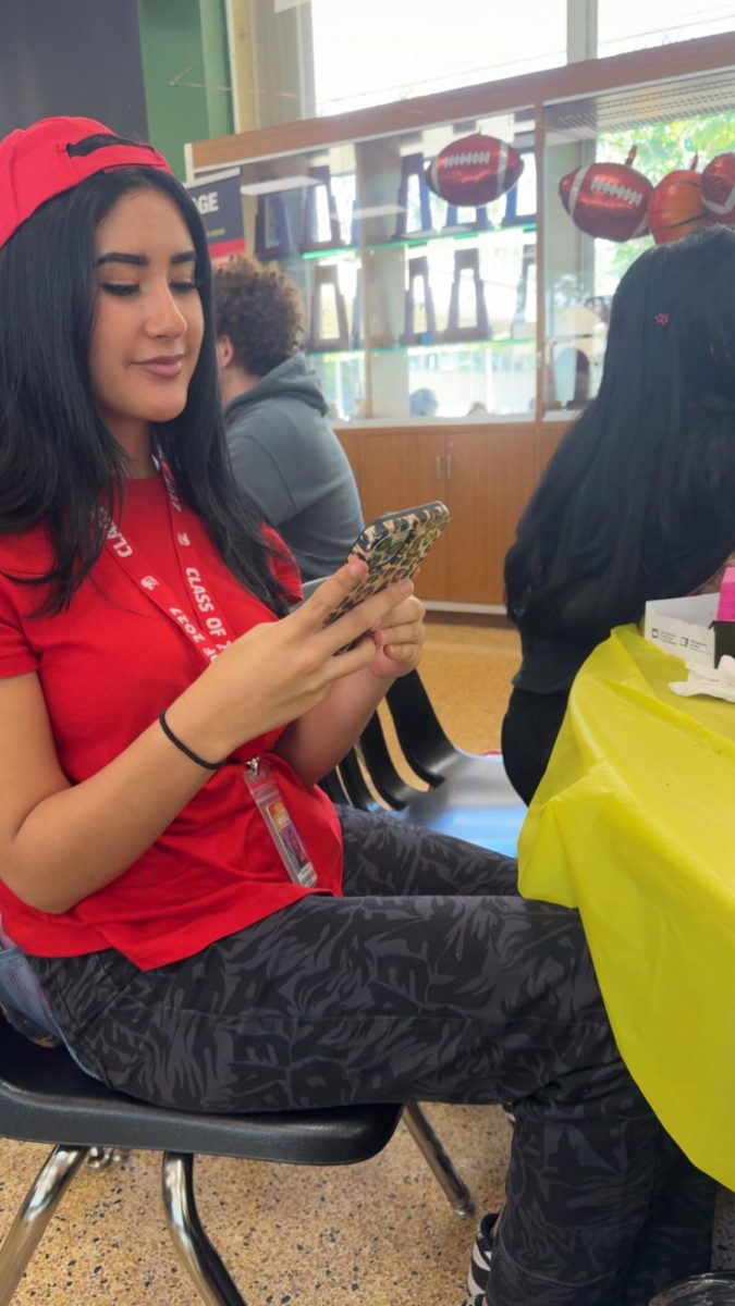 Senior Sofia Ferrer wears a red shirt and backward cap while checking her phone during lunch.