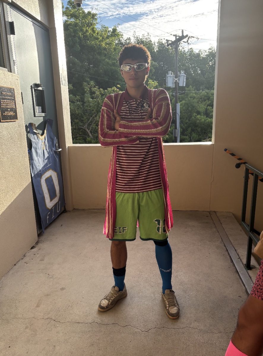 A student shows off his unique outfit with bright green shorts, striped clothing, and sunglasses for school spirit day.